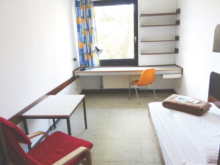 Zimmer Karl Egermann Haus Blick in ein klassisches Einzelzimemr mit Bett, Schreibtisch und Sessel